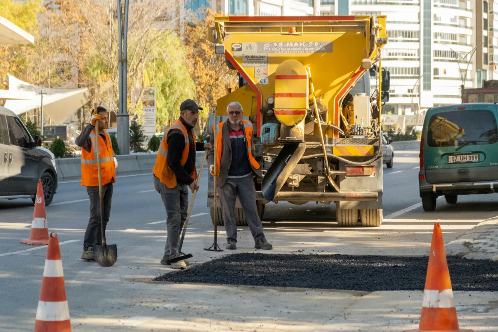 Road crew in safety vests preparing an asphalt patch repair beside traffic cones